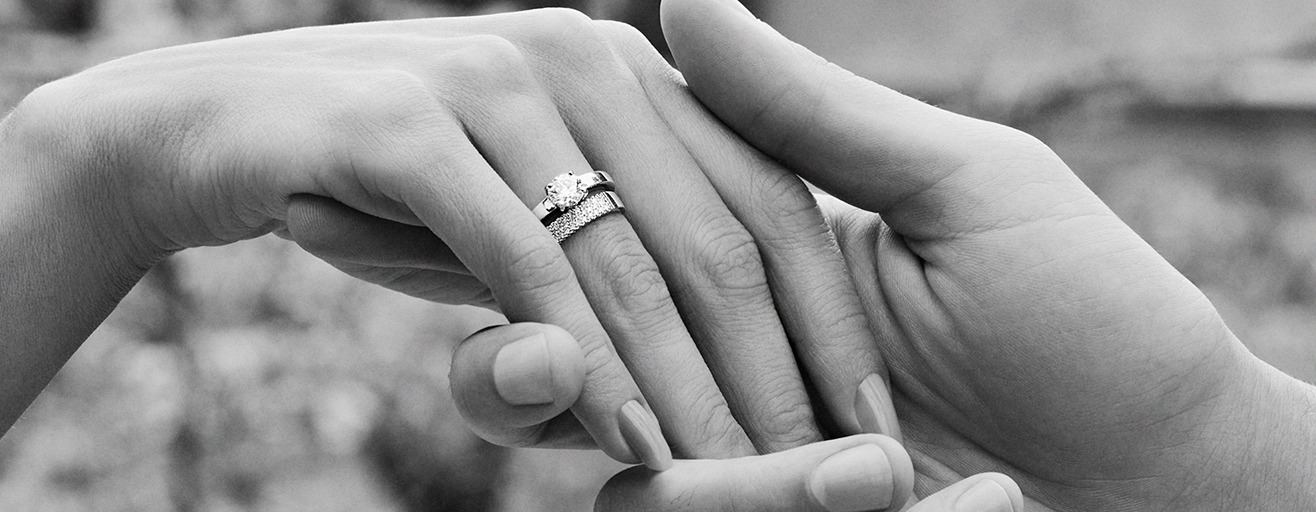 Na imagem é possível ver, em destaque, duas mãos. Em uma delas, existem 2 anéis prateados. A foto está em preto e branco.