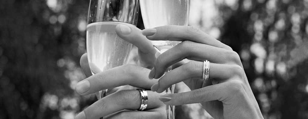Na imagem é possível ver duas mãos com alianças prateadas segurando taças de champagne. A foto está em preto e branco