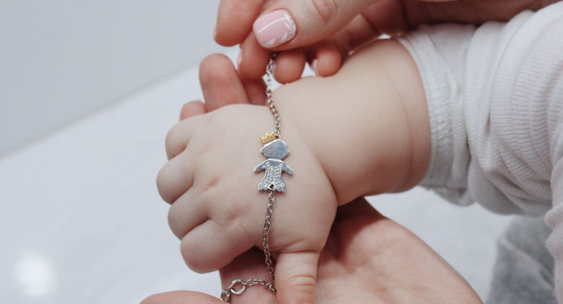 Na imagem é possível ver duas mãos femininas e 1 pequeno braço infantil segurando uma pulseira para bebê.