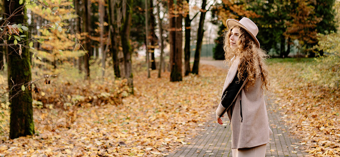 Mulher jovem, de cabelos claros e longos, usando uma roupa em tons pastel e um chapéu na mesma tonalidade, enquanto caminha por um parque com árvores e folhas caídas, em tons alaranjados de outono