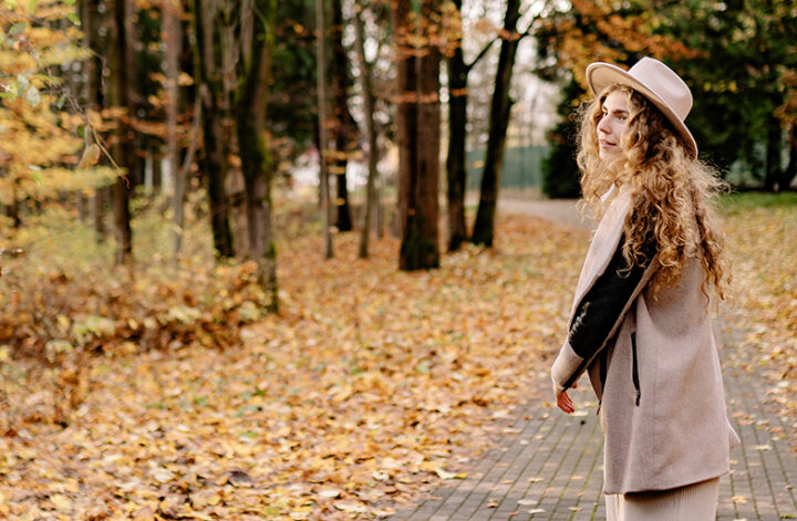 Mulher jovem, de cabelos claros e longos, usando uma roupa em tons pastel e um chapéu na mesma tonalidade, enquanto caminha por um parque com árvores e folhas caídas, em tons alaranjados de outono
