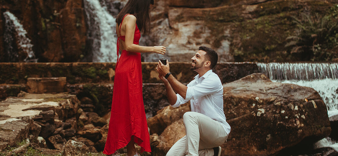 na imagem, um homem com roupas brancas pede uma mulher com vestido vermelho em casamento, de joelhos, com uma caixa de aliança na mão. Ao fundo, está uma cachoeira