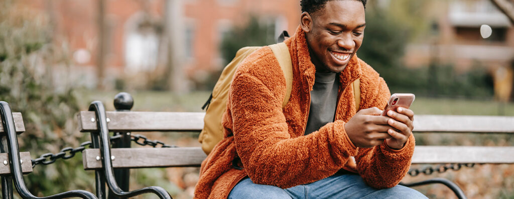 na imagem, um homem negro está sentado em um banco que se localiza em uma praça pública, enquanto veste um casaco e uma mochila em tons alaranjados e sorri, enquanto olha seu celular