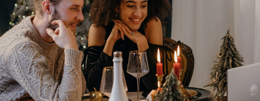na imagem, um casal está sentado à mesa, jantando, com velas acesas. Eles sorriem enquanto olham para um dispositivo eletrônico, como um tablet
