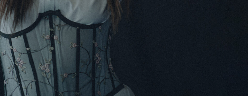 na imagem, a cintura da uma mulher está em foco: ela veste um corset transparente, na cintura, que contém imagens de flores delicadas e está sendo usado por cima de uma camisa branca