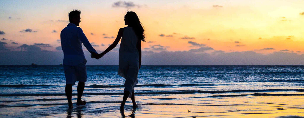na imagem, um casal está caminhando de mãos dadas à beira da praia, em um por do sol
