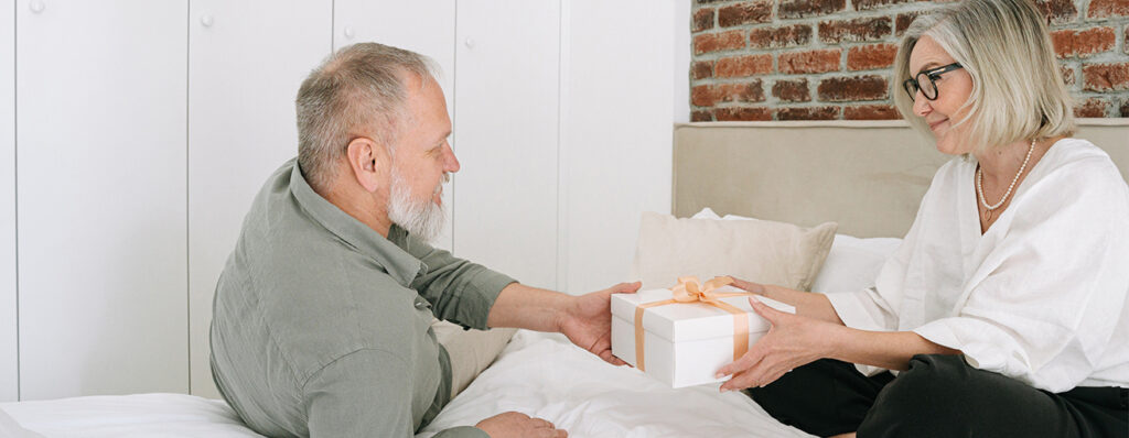 na imagem, um homem com camisa cinza está entregando um presente com laço salmão para uma mulher. Ambos estão em um ambiente interno de uma casa, sentados na cama