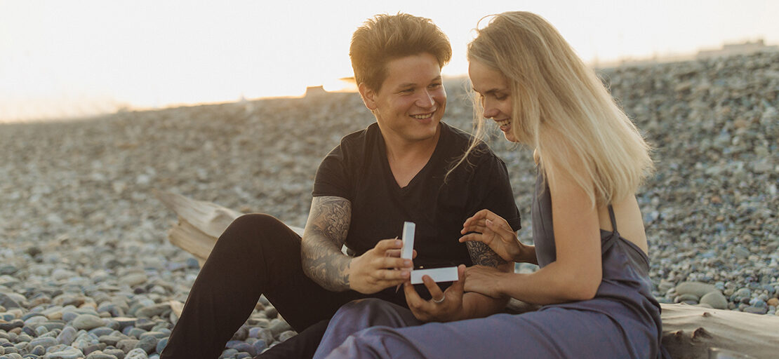 na imagem, um casal de namorados formado por um homem branco e loiro, com tatuagens, e uma mulher loira, estão sentados em um local aberto, como uma praia, ao por do sol, enquanto sorriem. O homem está segurando o que parece ser uma caixa de joias, entregando para a mulher