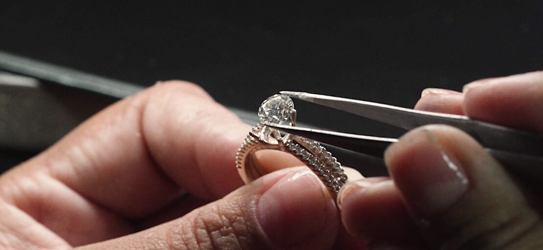 na imagem, duas mãos masculinas estão adicionando, com a ajuda de uma pinça, uma gema de Diamante ao aro de um anel solitário Vivara