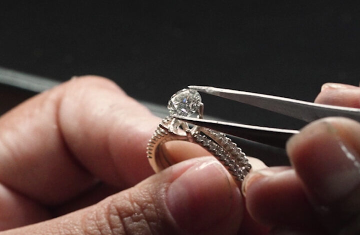 na imagem, duas mãos masculinas estão adicionando, com a ajuda de uma pinça, uma gema de Diamante ao aro de um anel solitário Vivara