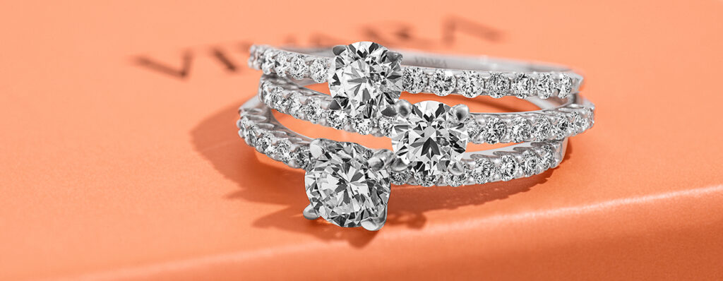 na imagem, estão três anéis Vivara com diamantes cravejados, dispostos sobre uma caixinha de presentes na cor salmão Vivara 