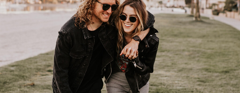na imagem, um casal está abraçado, rindo, enquanto caminha por um local aberto. Eles vestem roupas e óculos escuros.