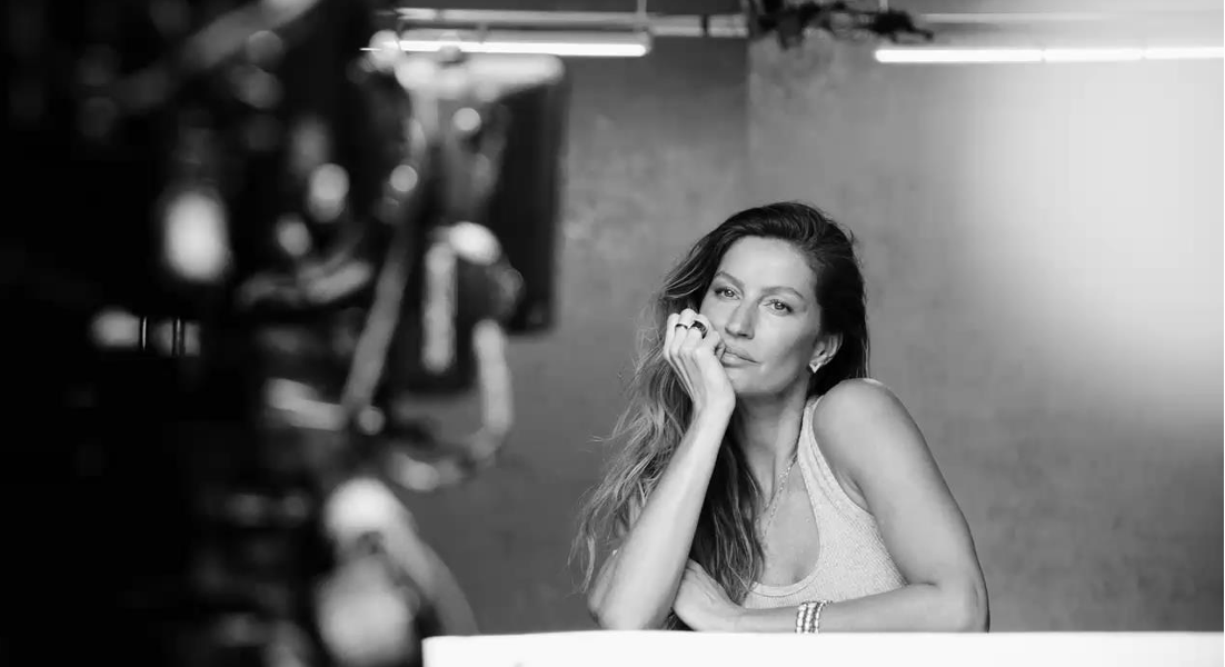 Imagem em preto e branco capturada durante o making of da campanha de Dia das Mães da Vivara. A Gisele Bündchen está sentada, com o rosto apoiado em uma das mãos, usando uma regata clara e joias delicadas, como brincos, anéis e pulseiras que fazem parte da nova coleção de joias especial Dia Das Mães 2025.