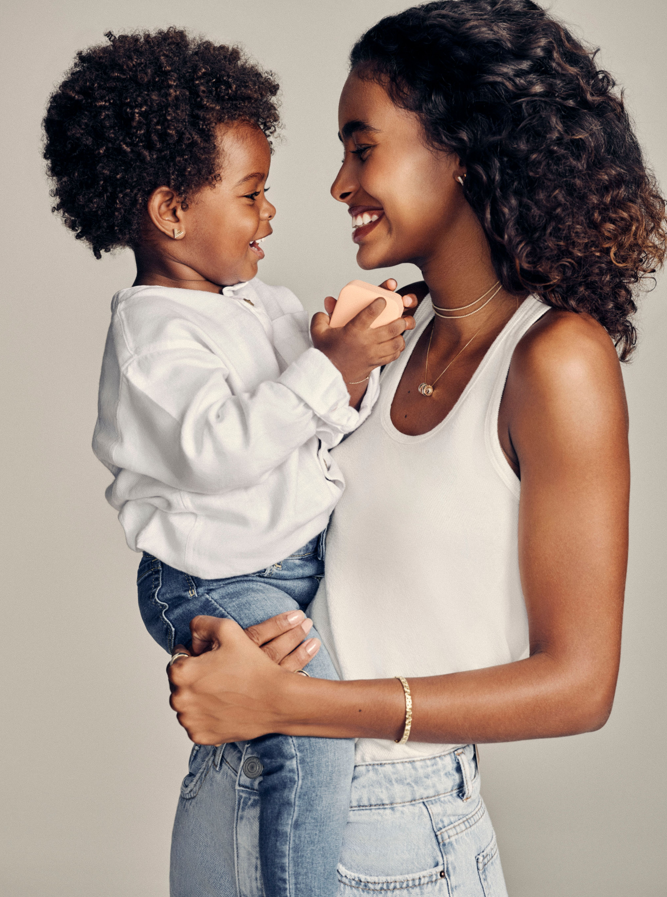 Mãe sorridente segurando sua filha no colo, ambos demonstrando carinho e felicidade. A mãe usa joias delicadas da coleção Life By Vivara, destacando o afeto do momento especial do Dia das Mães.