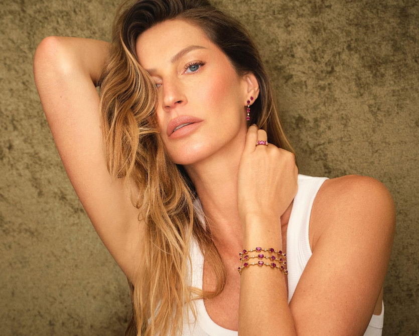 Imagem da Gisele Bündchen estrelando a coleção de joias Duquesa. Ela veste brincos, pulseiras e anel em tons de rosa vibrante, com design sofisticado e detalhes delicados. Ela apoia uma das mãos no pescoço e exibe uma pose elegante, vestindo uma blusa branca simples que destaca as joias.