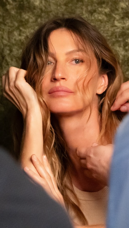 Gisele Bündchen, com expressão serena, é preparada para a sessão de fotos da campanha de Dia das Mães Vivara 2025, com os cabelos sendo ajustados pelos profissionais de beleza.